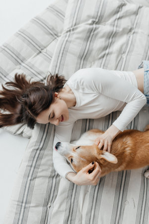 In a serene indoor setting, a young woman cherishes a delightful and joyful moment spent with her playful corgi while sitting on a cozy blanket, radiating warmth and happinessの写真素材