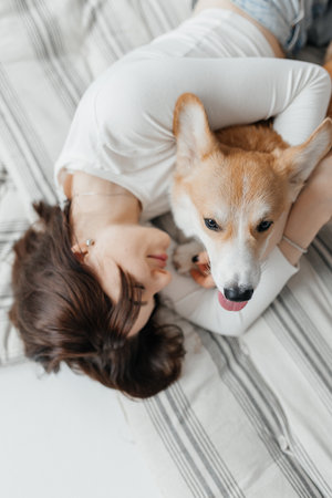 A woman relaxes on her cozy bed, snuggling with her affectionate Corgi. This scene highlights their loving bond, creating a warm, joyful atmosphere in their homeの写真素材