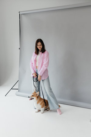 In a contemporary studio setting, a fashionable young woman presents a relaxed and chic vibe next to her adorable corgi, both styled against a soft, elegant gray backdropの写真素材