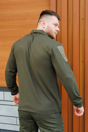 A male model displays an olive green outdoor jacket, striking a confident pose in front of rustic wooden panels, making it ideal for outdoor fashion photoshootsの写真素材