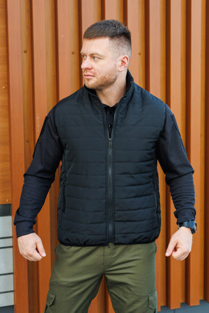 A confident man poses in a sleek black puffer vest and a stylish outfit against a modern urban backdrop, showcasing the latest fashion trendsの写真素材