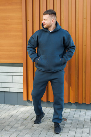 A young man confidently poses against a wooden wall, dressed in a trendy dark blue hoodie paired with comfy jogging pants, epitomizing the essence of modern casual fashionの写真素材