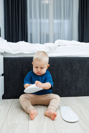 Cute blond toddler boy in blue t-shirt running in spacious hotel room. Toddler boy in comfortable hotel room resting after travel. Child at home sitting on floorの写真素材