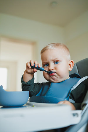 Smiling boy eats soup sitting on a chair. Hungry boy puts food in his mouth. Baby one year and three months old eats independently. Silicone utensils for feeding children.の写真素材