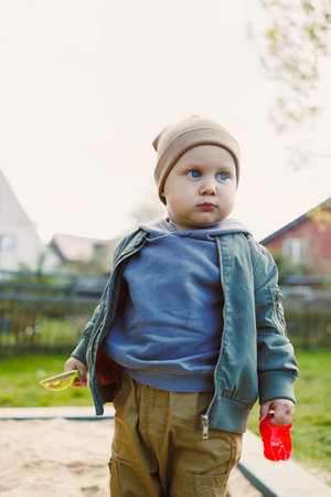 A boy of 2 years old in a spring jacket and hat plays with sand in a sandbox. A boy plays on a...