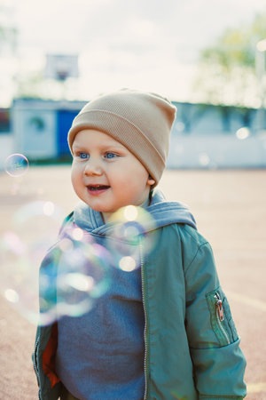 Toddler boy running outside with soap bubbles. Child on a warm spring day for a walk in a jacket and...