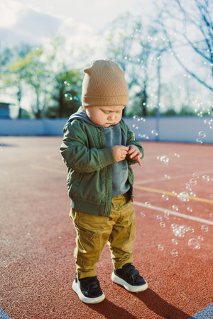 Cheerful 2 year old boy playing outside in summer in a green jacket with bubbles. Toddler boy...