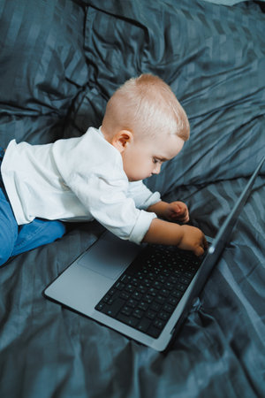 Cute boy 2 years old sits in bed and presses keys on laptop. Child sits and watches video on laptop....