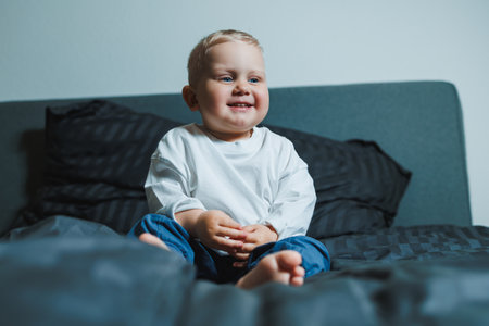 Cute toddler boy in white shirt and pants laughing while sitting in bed. 2 year old boy playing while sitting in bed. Home clothes for boys.の写真素材