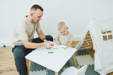 Dad teaches his 1.6 year old son to sculpt from plasticine. The toddler boy sits at the children's table with his dad and plays entertaining games. The child sculpts figures from plasticineの写真素材