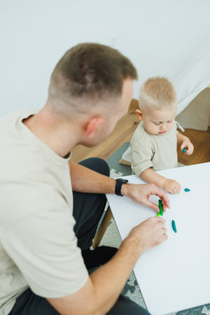 Dad teaches his 1.6 year old son to sculpt from plasticine. The toddler boy sits at the children's table with his dad and plays entertaining games. The child sculpts figures from plasticineの写真素材