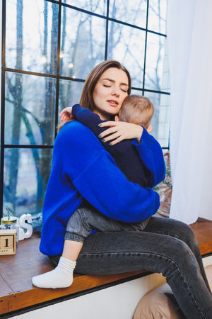 Happy mother and her 4 year old son hugging while sitting on the windowsill at home. Trusting relationship between mother and son. Little boy hugging his mother.の写真素材
