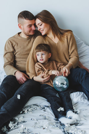 A loving couple joyfully interacts with their adorable child on a cozy bed decorated with silver confetti, capturing heartwarming family memories and togethernessの写真素材