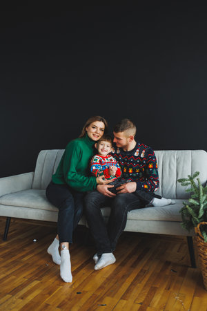 A joyful family portrait featuring two loving parents and their adorable child in vibrant holiday sweaters, smiling on a cozy couch, embodying happiness during the festive seasonの写真素材