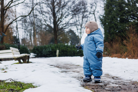 In a beautifully snowy park, a joyful toddler dressed in a bright blue snow suit plays happily among the tall trees, truly embodying pure childhood winter fun and laughterの写真素材