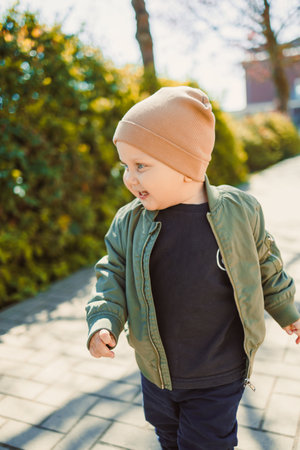 A cheerful child is enjoying a sunny day on a pathway, wearing a trendy bomber jacket and cozy hat,...