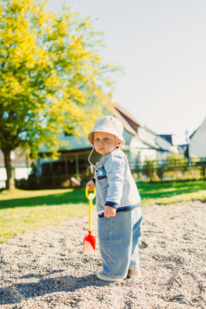 On a beautiful sunny day, a cheerful young child happily explores a sandy area with a brightly colored shovel, surrounded by lush greenery, trees, and playground equipmentの写真素材