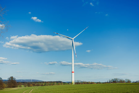 A tall wind turbine captures wind energy in a green field against a blue sky, symbolizing a shift towards sustainable energy solutions that benefit our environmentの写真素材