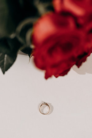A stunning and exquisite golden ring gracefully rests on a pristine white surface, beautifully framed by vibrant red roses, symbolizing love, deep commitment, and timeless romanceの写真素材