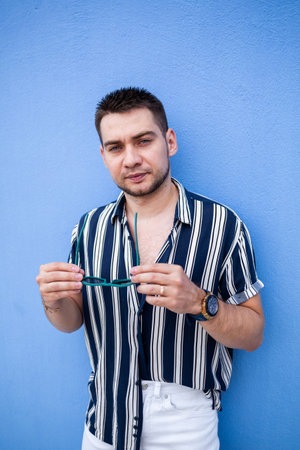 A fashionable young man stands confidently against a blue background, holding sunglasses and showcasing a trendy summer look with a striped shirt and white pants.の写真素材