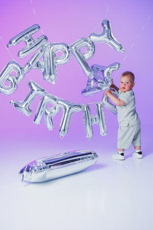A cheerful toddler plays with shiny silver Happy Birthday balloons against a colorful backdrop, ideal for joyful birthday celebrations with family and friendsの写真素材