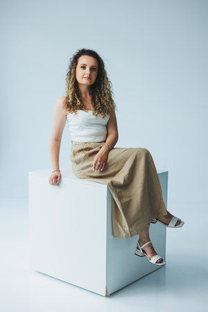 Curly woman sitting on a white box and smiling dreamily. Woman in a white top and skirt with a romantic mood sitting on a white box.の写真素材