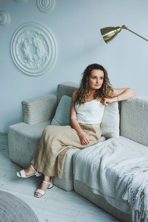 Curly pretty woman sitting in the living room on the sofa and smiling dreamily. Woman in a white top and skirt with a romantic mood sitting in the living room.の写真素材