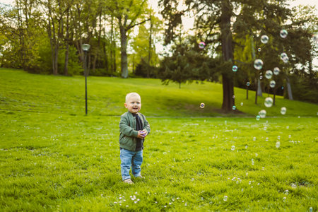 Little 2 year old boy running on green grass with soap bubbles. Toddler boy enjoying a walk in nature. Child having fun in city park.の写真素材