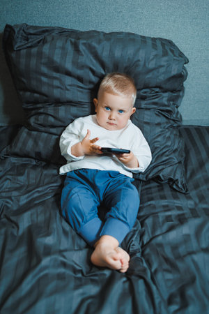 Boy holds phone in hands. Toddler boy in white t-shirt and jeans sits on bed and watches cartoons on phone. 2 year old boy plays with mobile gadget. Modern digitalization of kids.の写真素材