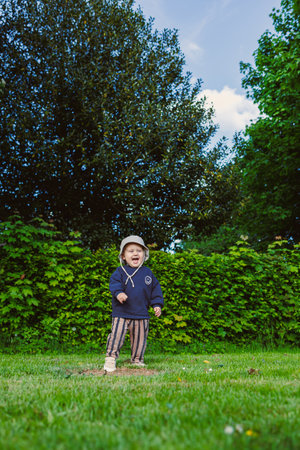 Smiling toddler boy in a panama hat on his head runs on green grass in a city park. A walk of a 2 year old boy in nature. The child enjoys the beauty of nature and a sunny day in summer.の写真素材