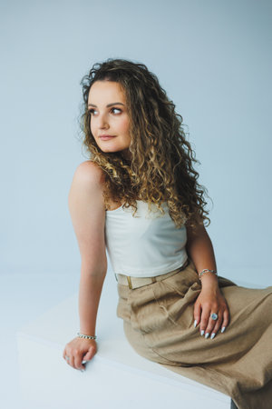 A young woman with beautiful curly hair, dressed in a stylish outfit, sits gracefully in a minimalist studio, exuding confidence and showcasing her chic sense of fashionの写真素材