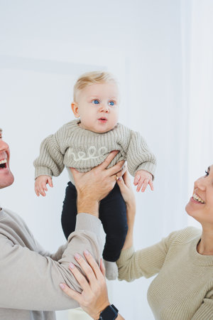 In a warm home filled with love, a cheerful family gathers around, joyfully lifting their baby,...