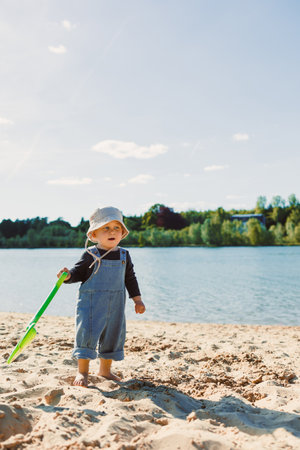 Toddler in a panama hat on his head and with bare feet walks on the sand on the beach. 2 year old boy digs golden sand on a warm sunny day. Child on a walk on the beach.の写真素材