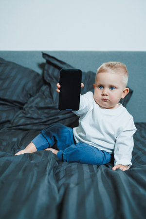 Handsome toddler boy in a white t-shirt and jeans sits on the bed and watches cartoons on the phone. 2 year old boy plays with a mobile gadget. Modern digitalization of toddlers.の写真素材
