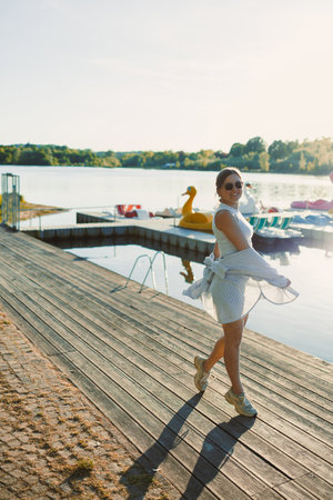 Cheerful woman in shorts and sunglasses walking along the pier near the lake. Woman having fun and running under the scorching sun. Young woman on a walk near the lake.の写真素材