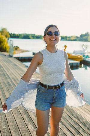 Cheerful woman in shorts and sunglasses walking along the pier near the lake. Woman having fun and running under the scorching sun. Young woman on a walk near the lake.の写真素材