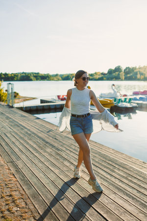 Cheerful woman in shorts and sunglasses walking along the pier near the lake. Woman having fun and running under the scorching sun. Young woman on a walk near the lake.の写真素材
