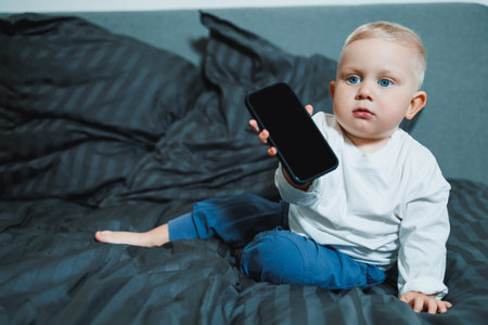 Handsome toddler boy in a white t-shirt and jeans sits on the bed and watches cartoons on the phone. 2 year old boy plays with a mobile gadget.の写真素材