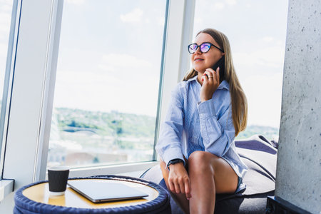 A woman is sitting comfortably in front of a large window, engaged in an animated conversation on...
