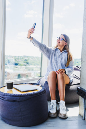 A woman is comfortably sitting on a soft bean bag chair while she takes a selfie using her...