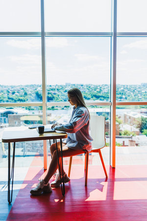A woman is seated comfortably at a desk that is positioned in front of a large window, allowing natural light to fill the spaceの写真素材