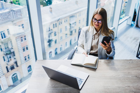 A woman is comfortably seated at her desk, engaging with a laptop and using her cell phone for various tasks and communicationsの写真素材