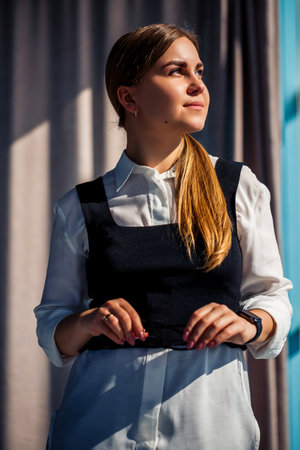 A poised young woman stands elegantly in stylish attire, illuminated by natural light, exuding sophistication, individuality, and creativity in a modern settingの写真素材