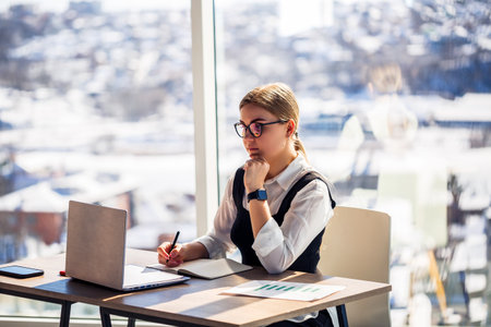 A woman with glasses sits at a modern desk, focused on her laptop, with a beautiful snowy landscape visible through large windows, creating a serene atmosphereの写真素材