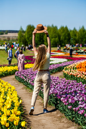 A happy young woman with long, flowing hair joyfully raises her stylish hat while immersed in the stunning beauty of a vibrant tulip field on a warm and sunny spring dayの写真素材