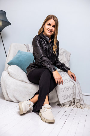 A fashionable young woman sits confidently on a cozy sofa, dressed in a sleek leather jacket and casual attire. A warm atmosphere is created with decorative elements.の写真素材