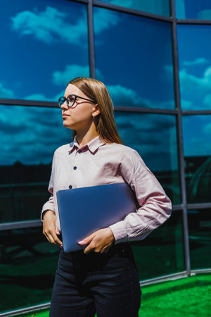 A confident young woman stands by large windows with her laptop, showcasing her professionalism and motivation, fueled by her ambition as she advances steadily in her careerの写真素材