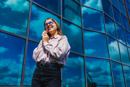 A fashionable young woman stands confidently against a reflective glass building, showcasing her style and positivity on a sunny day. Perfect for lifestyle themes.の写真素材