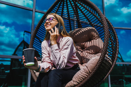 A stylish young woman relaxes in a cozy hanging chair, savoring coffee and chatting on her phone, enjoying a delightful moment under a clear blue sky, surrounded by natureの写真素材