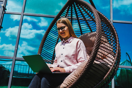 In a serene outdoor setting, a young woman is focused on her laptop while lounging in a cozy hanging chair, nurturing her creativity and enhancing productivityの写真素材